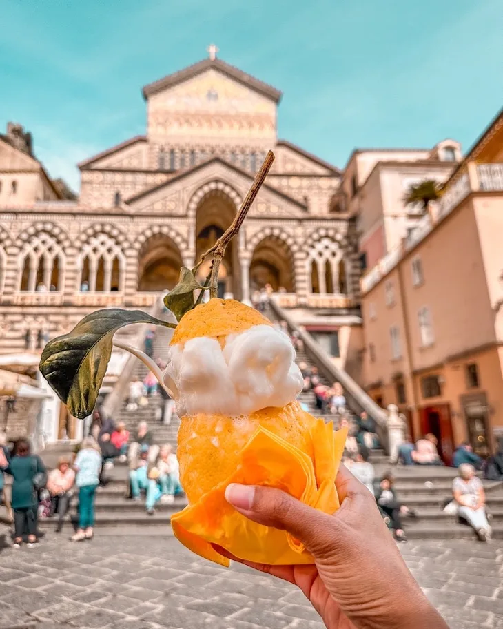 Hand holding a lemon-shaped gelato with a leaf in front of the striped cathedral steps and crowd in Amalfi.