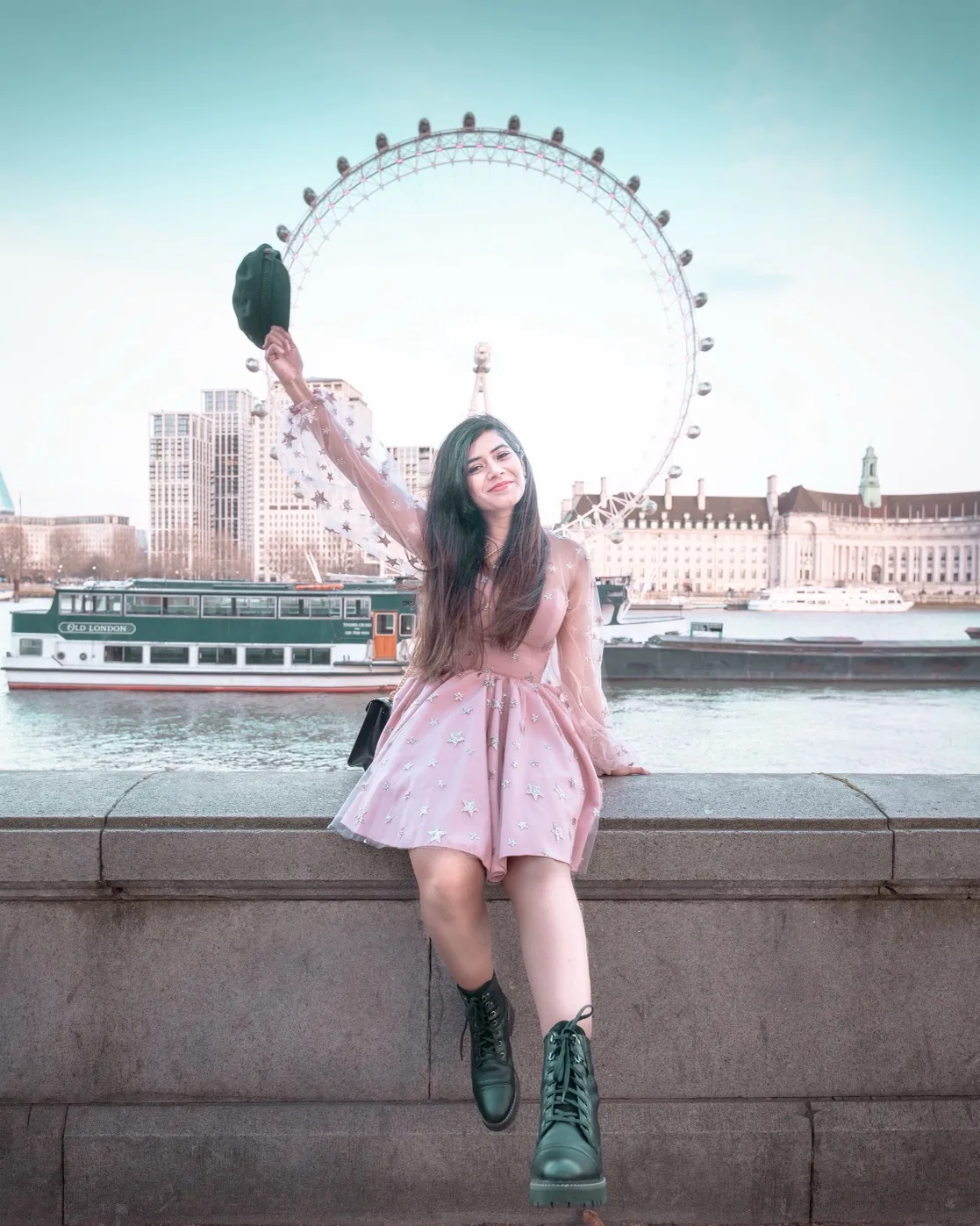 Young woman in a pink star-patterned dress and green boots sitting on a riverside wall, waving a hat with the London Eye and riverboats behind her.