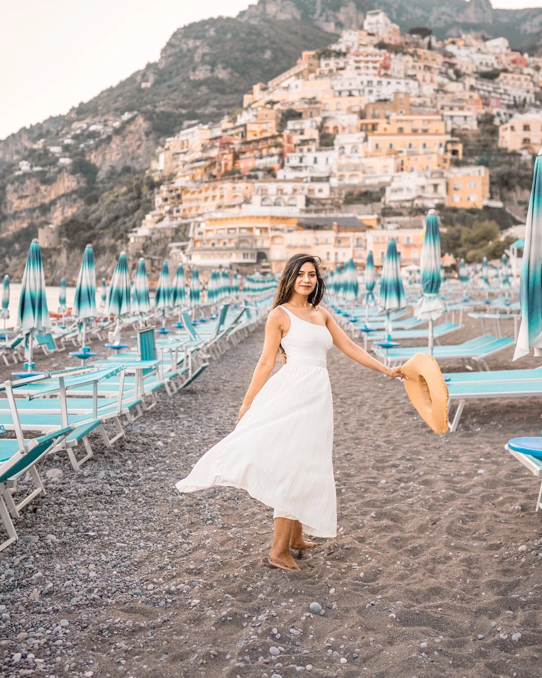 Woman in a white summer dress holding a straw hat on a pebble beach lined with turquoise umbrellas and sunbeds below cliffside pastel buildings.