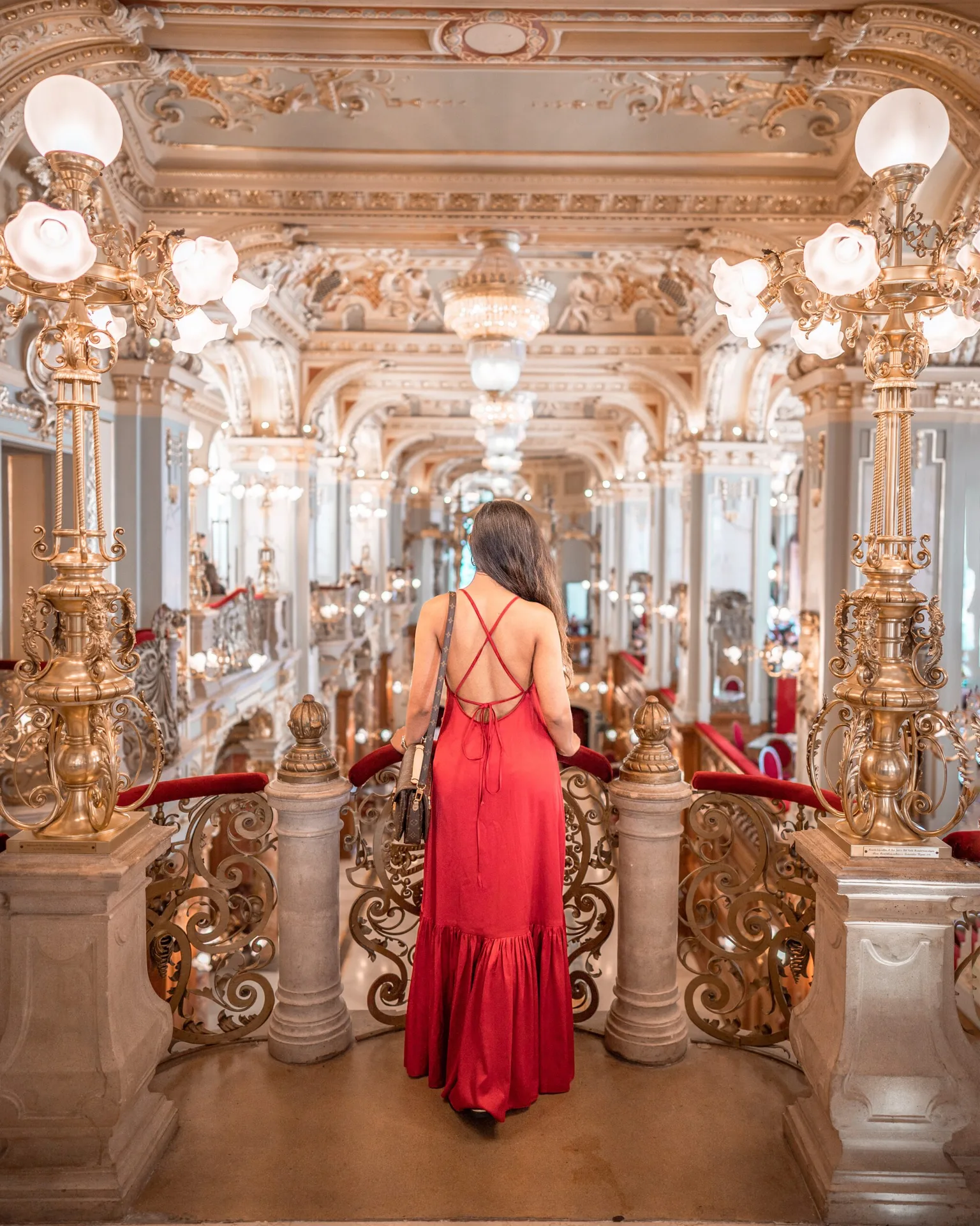 Woman in a red backless dress overlooking an ornate, gold‑filled historic ballroom from an upper balcony.