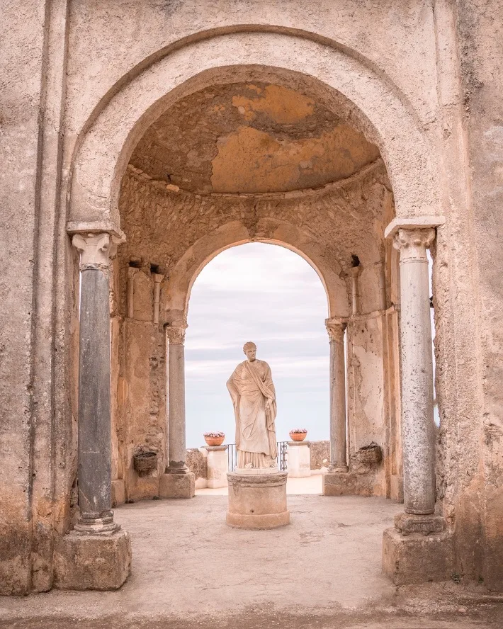 Marble statue of a robed figure framed by a weathered stone archway and classical columns overlooking a pale sky and distant sea.