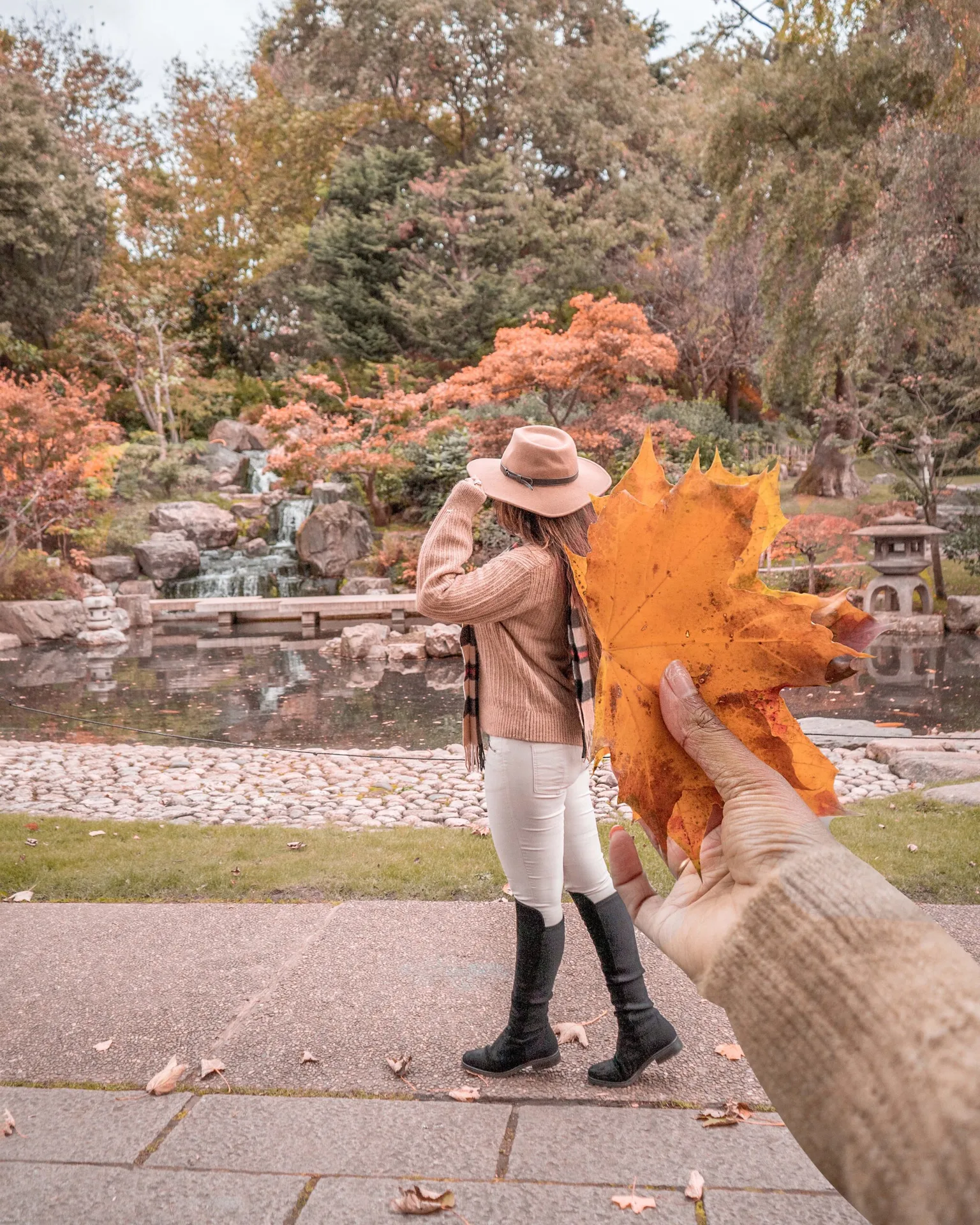 Person in a hat and autumn coat walks past a tranquil pond and waterfall while a hand holds a large orange maple leaf in the foreground.