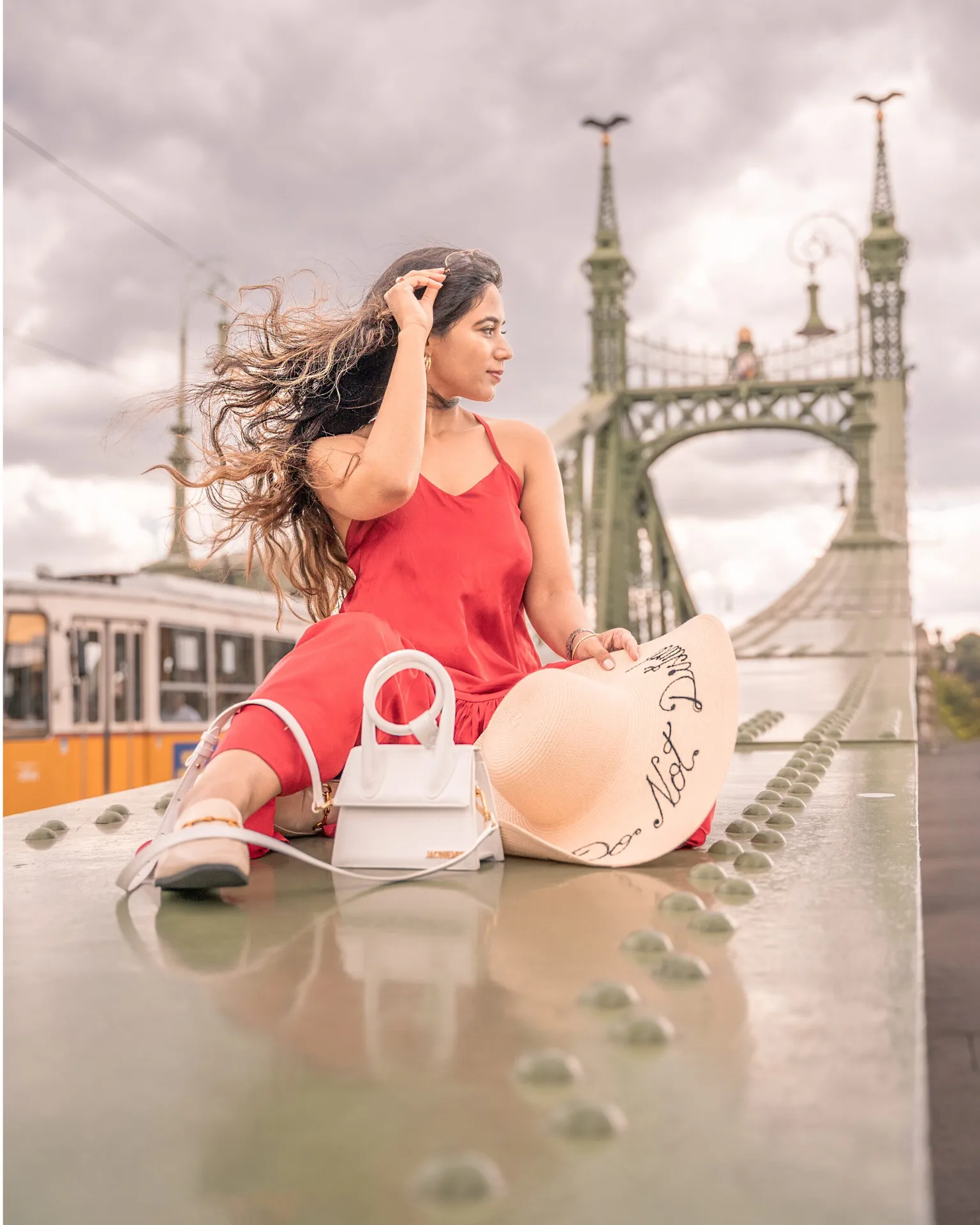 Woman in a red dress sits on a green ornate bridge with her hat and white handbag, hair blowing as a yellow tram passes behind.