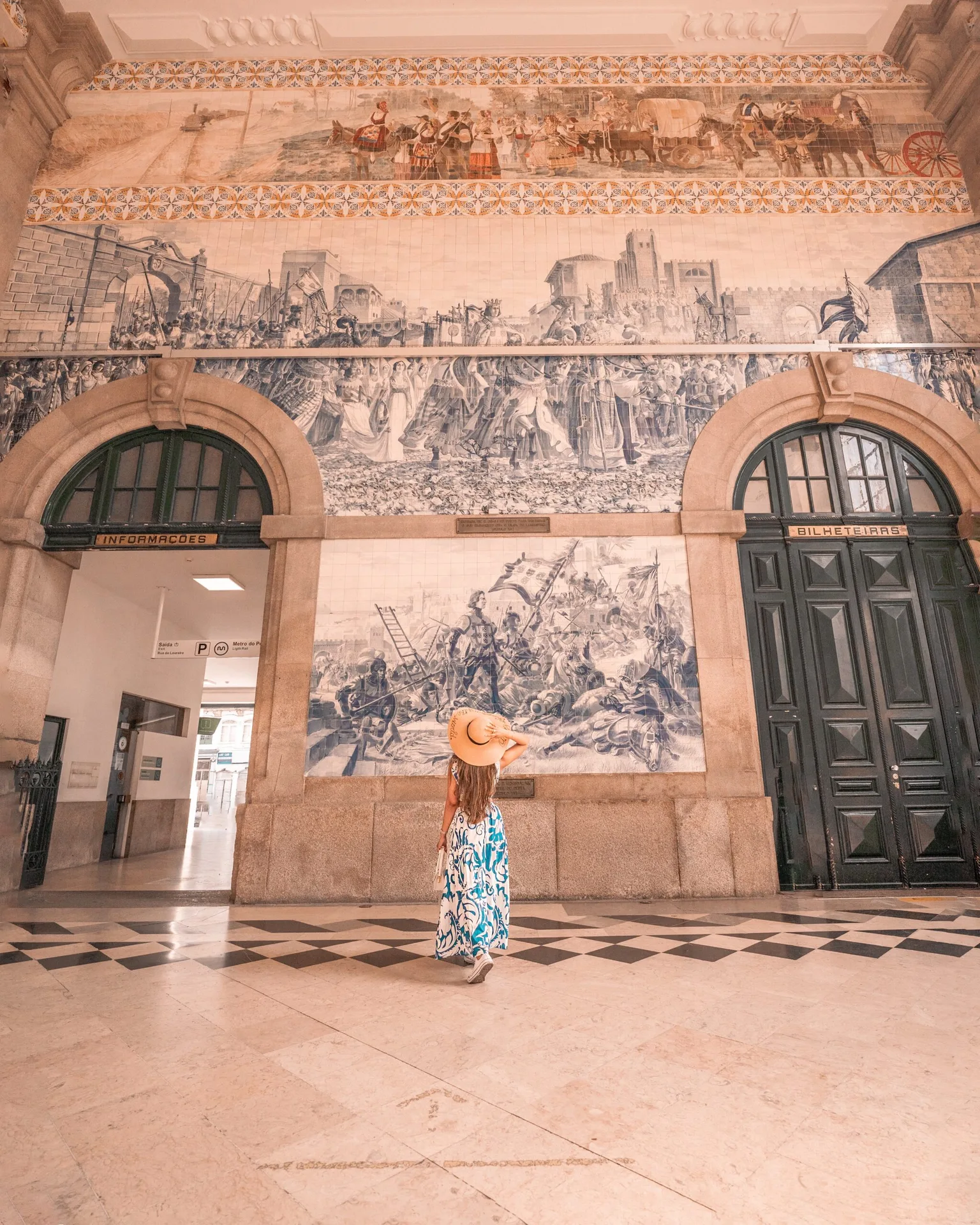 Woman in a sunhat and patterned dress stands in a tiled train station hall, admiring large historic blue-and-white azulejo murals above arched doorways.