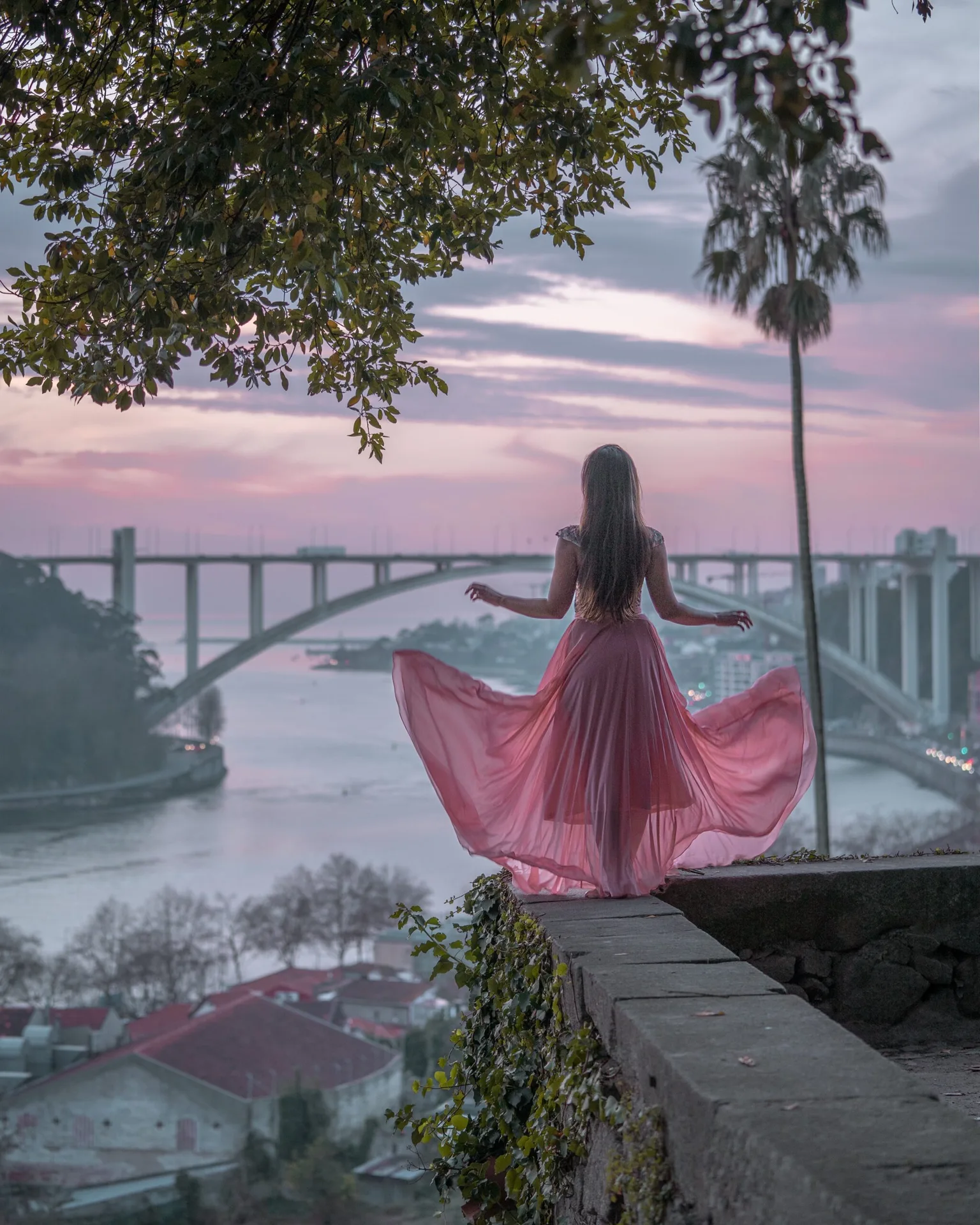 Woman in a flowing pink dress stands on a stone ledge overlooking a river and arched bridge at pastel sunset.