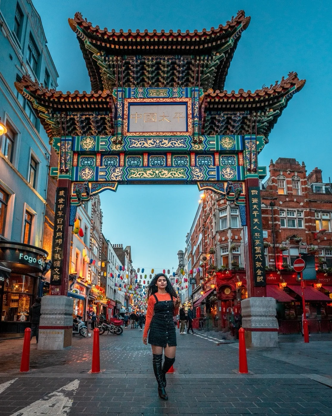 Woman in a black pinafore and orange top walking under a colourful ornate Chinese gate in a bustling lantern‑lined Chinatown street.