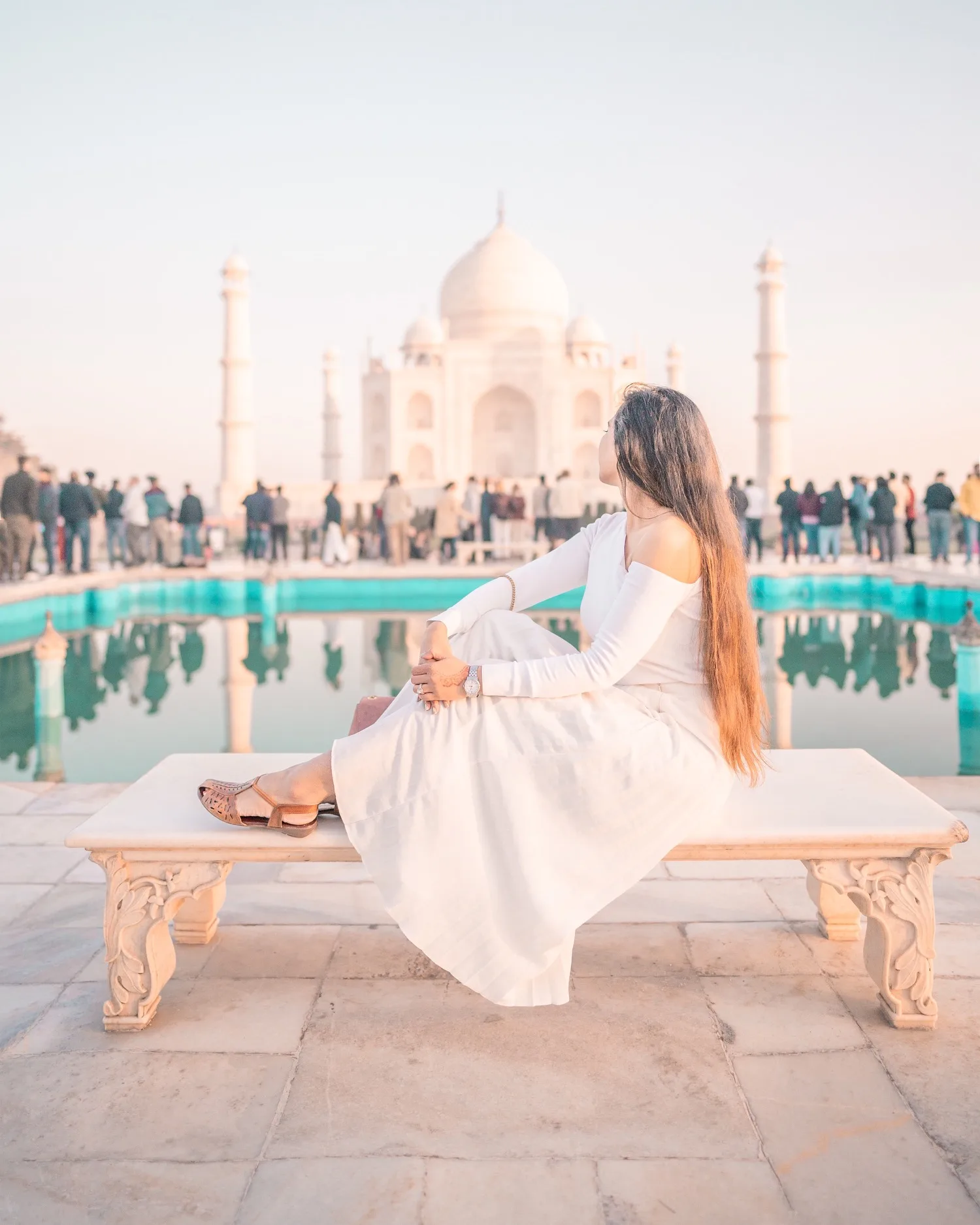 Woman in a white dress sitting on a marble bench gazing at the Taj Mahal reflected in the pool.