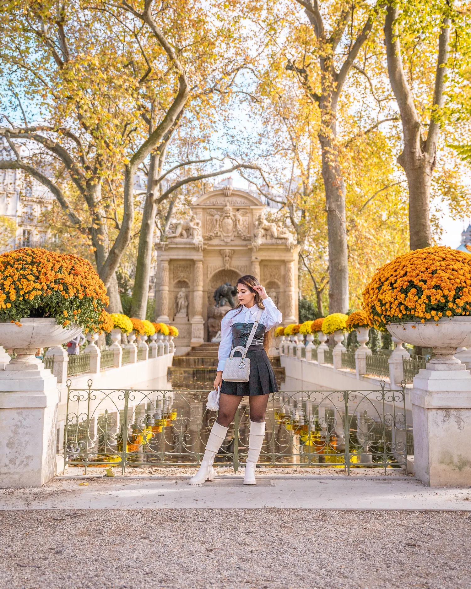 Young woman in a chic autumn outfit poses by a fountain and symmetrical orange mums in a sunlit Parisian garden.