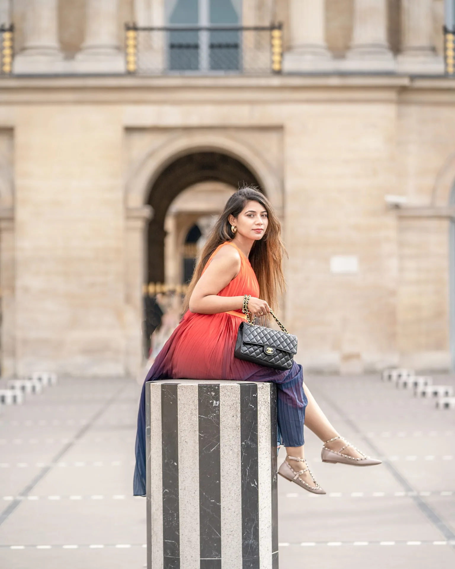 Woman in a flowing red-to-blue dress sitting on a striped marble column, holding a black quilted handbag with historic archways blurred behind.