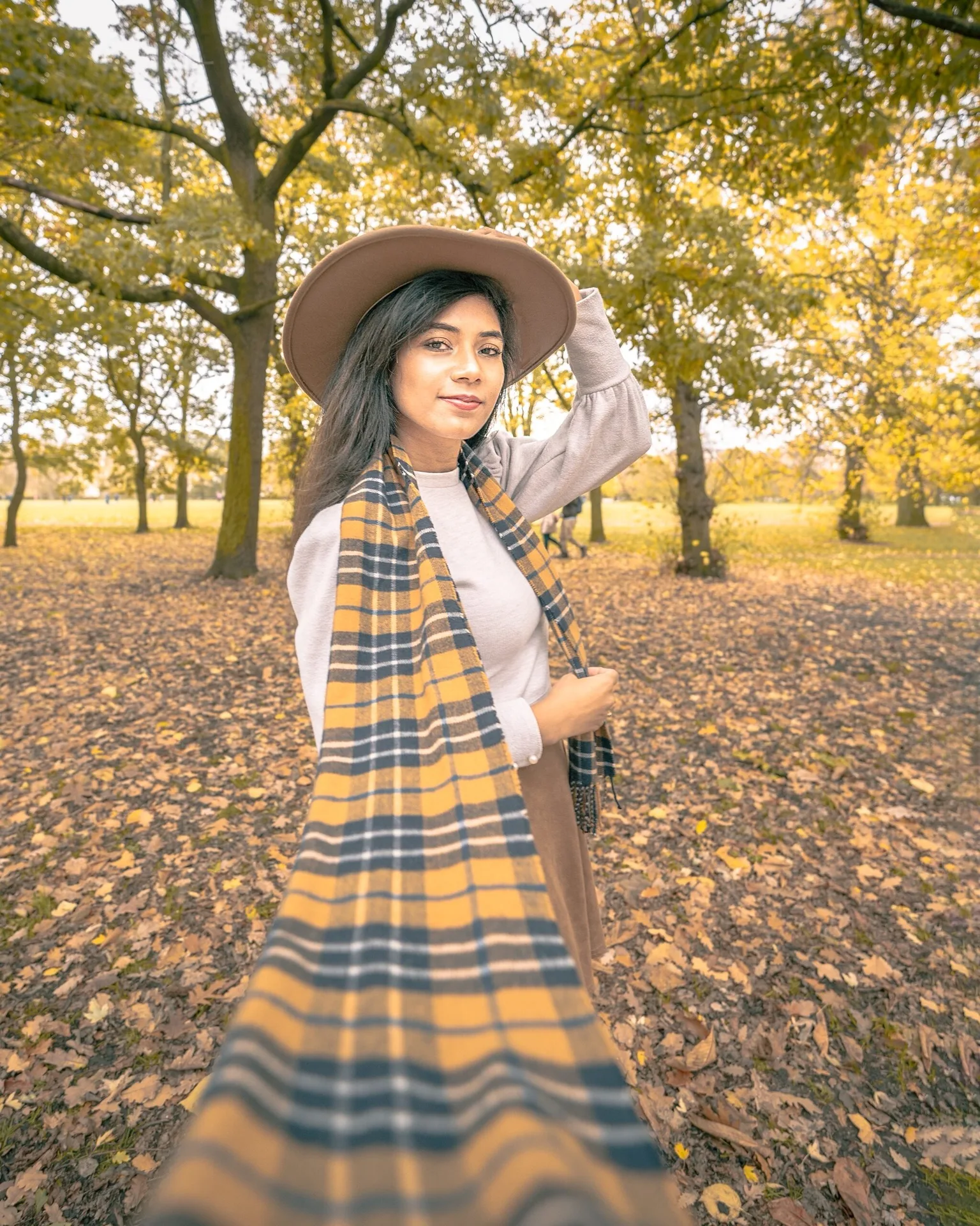 Young woman in a wide-brim hat and yellow plaid scarf holding out the camera while posing in a leafy autumn park.