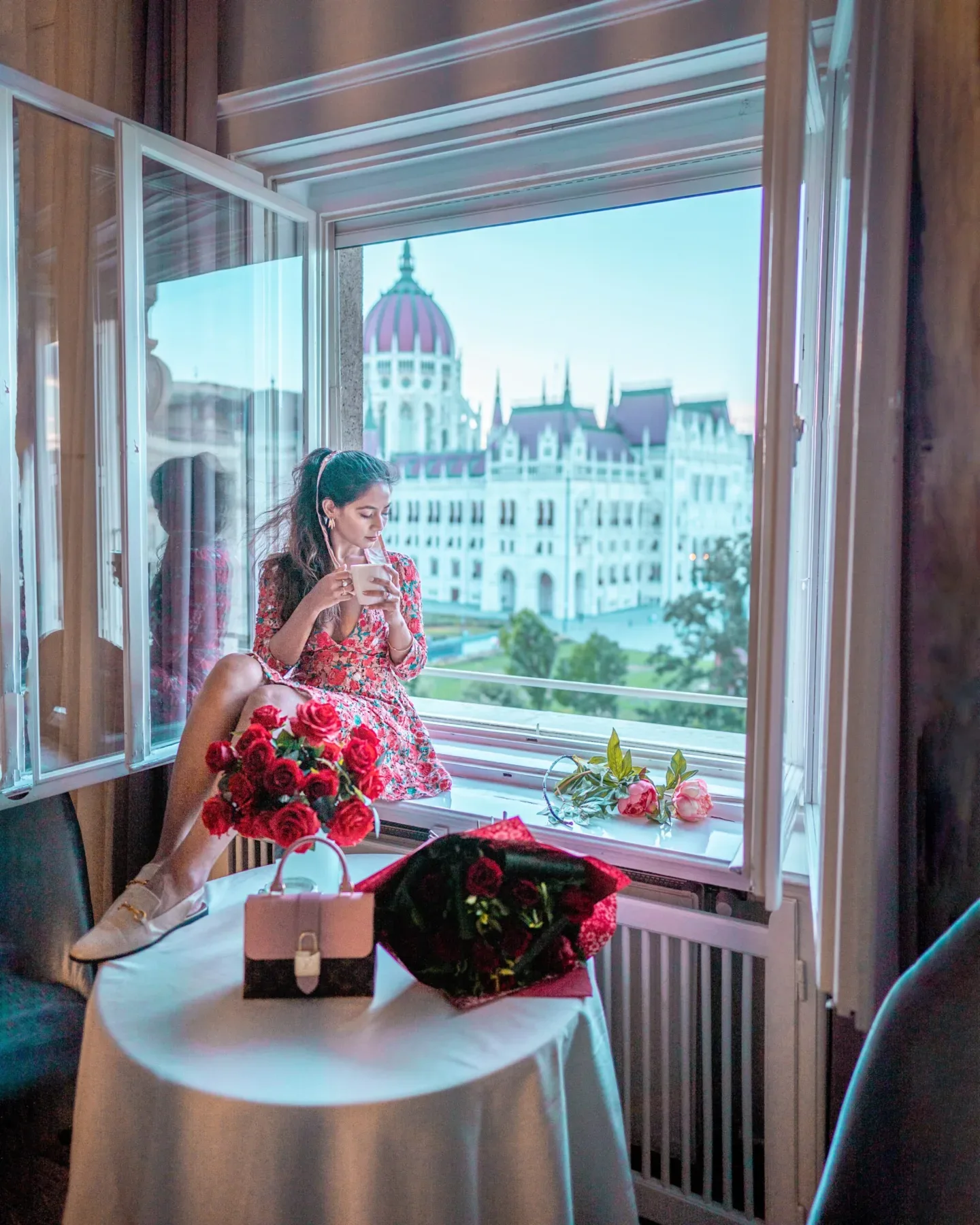 Woman in a floral dress sips a drink on a hotel windowsill overlooking the grand domed parliament building.