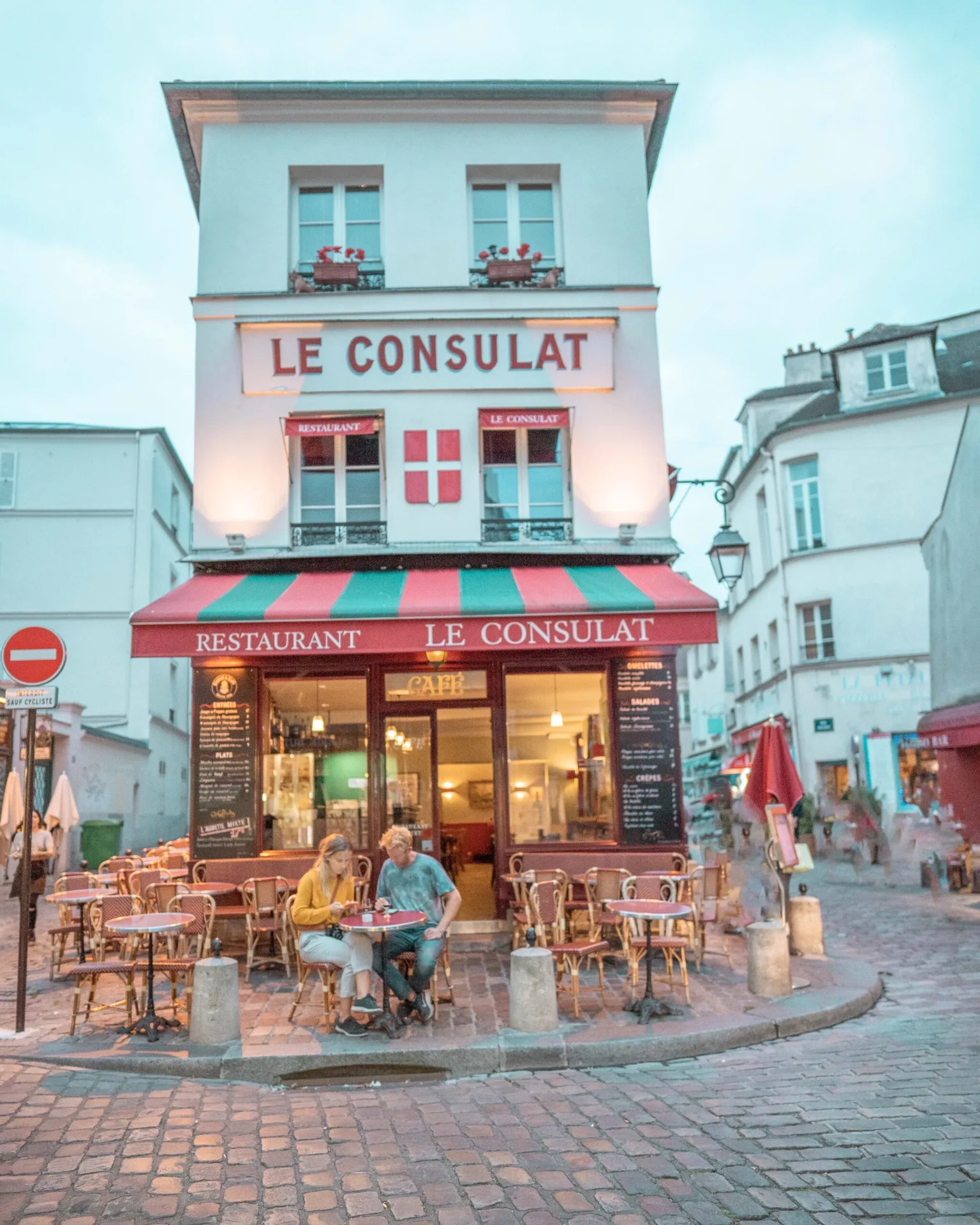 Couple sitting at a small table outside the classic red-and-green awned Le Consulat café on a cobbled Parisian street.