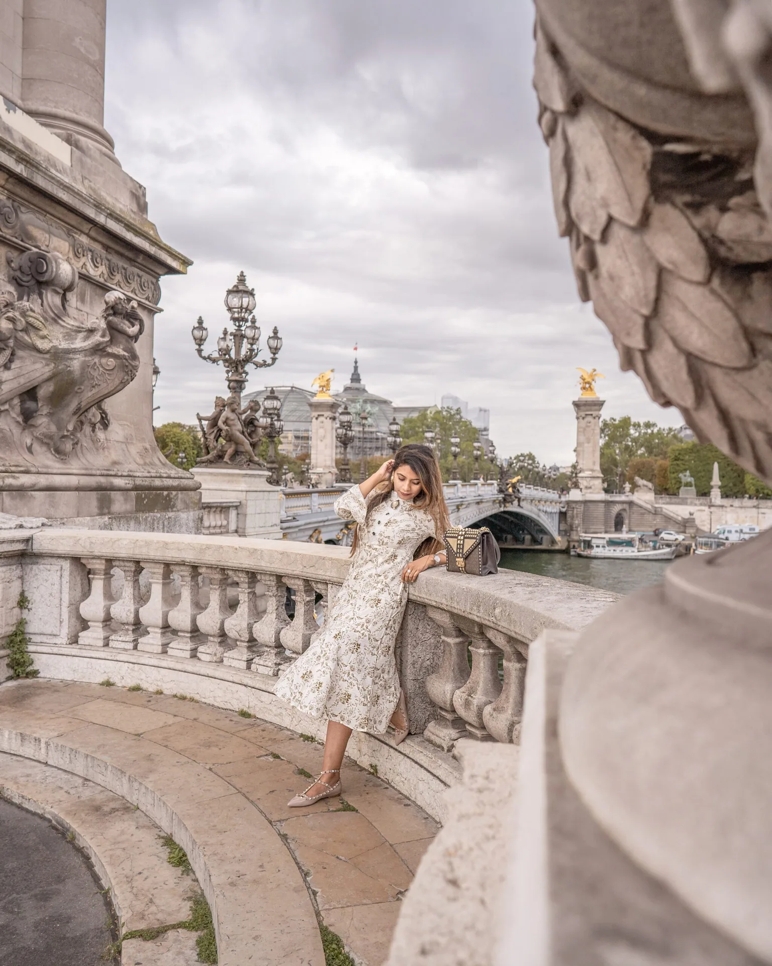 Woman in a floral dress leaning on the ornate stone balustrade of Pont Alexandre III with the Seine and gilded lampposts in the background.