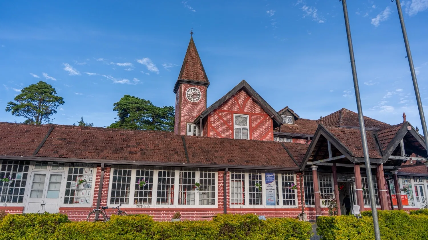 Red brick colonial-style train station with a tall clock tower, tiled roofs and white-framed windows under a clear blue sky.