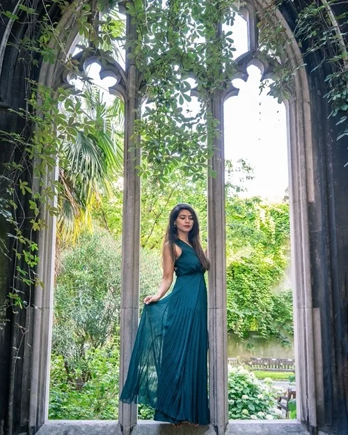 Woman in a flowing teal dress standing in a Gothic stone window frame overlooking a sunlit, vine-covered garden.