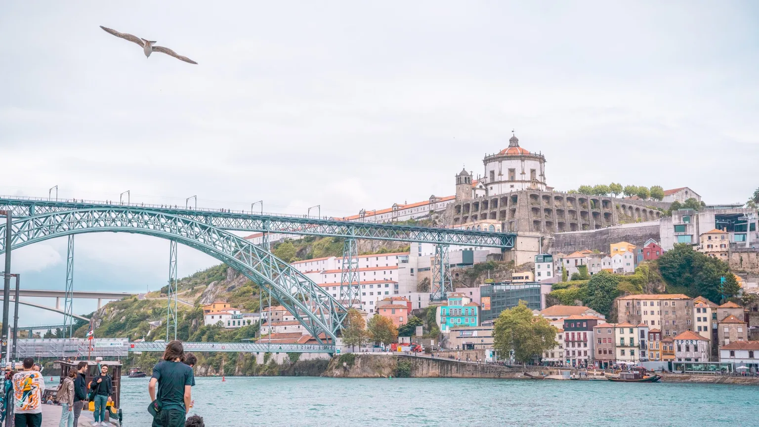 People strolling the riverside with the arched Dom Luís I Bridge and hillside buildings of Porto under a pale sky.