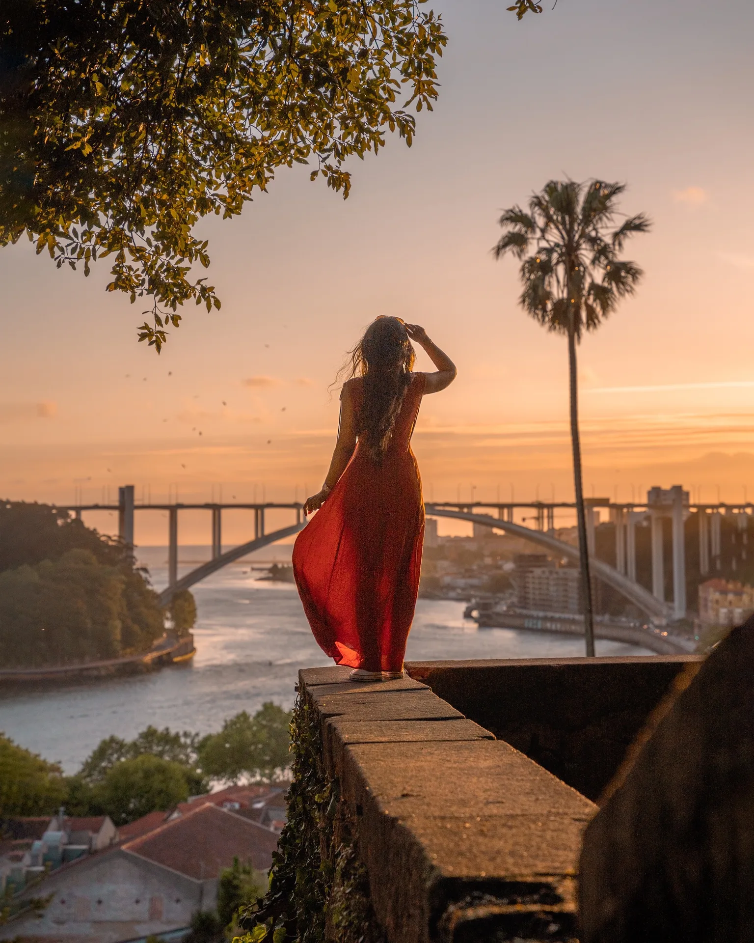 Woman in a flowing red dress standing on a stone ledge overlooking a river and arched bridge at golden sunset.