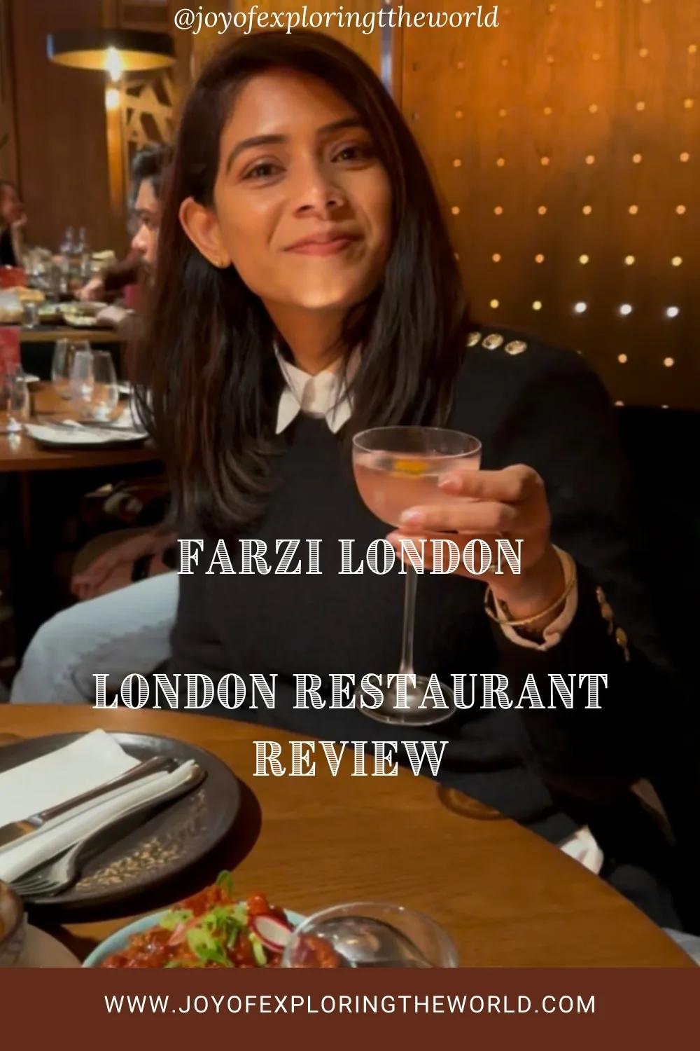 Woman smiling and raising a cocktail glass while seated at a warmly lit London restaurant table with plates and dishes nearby