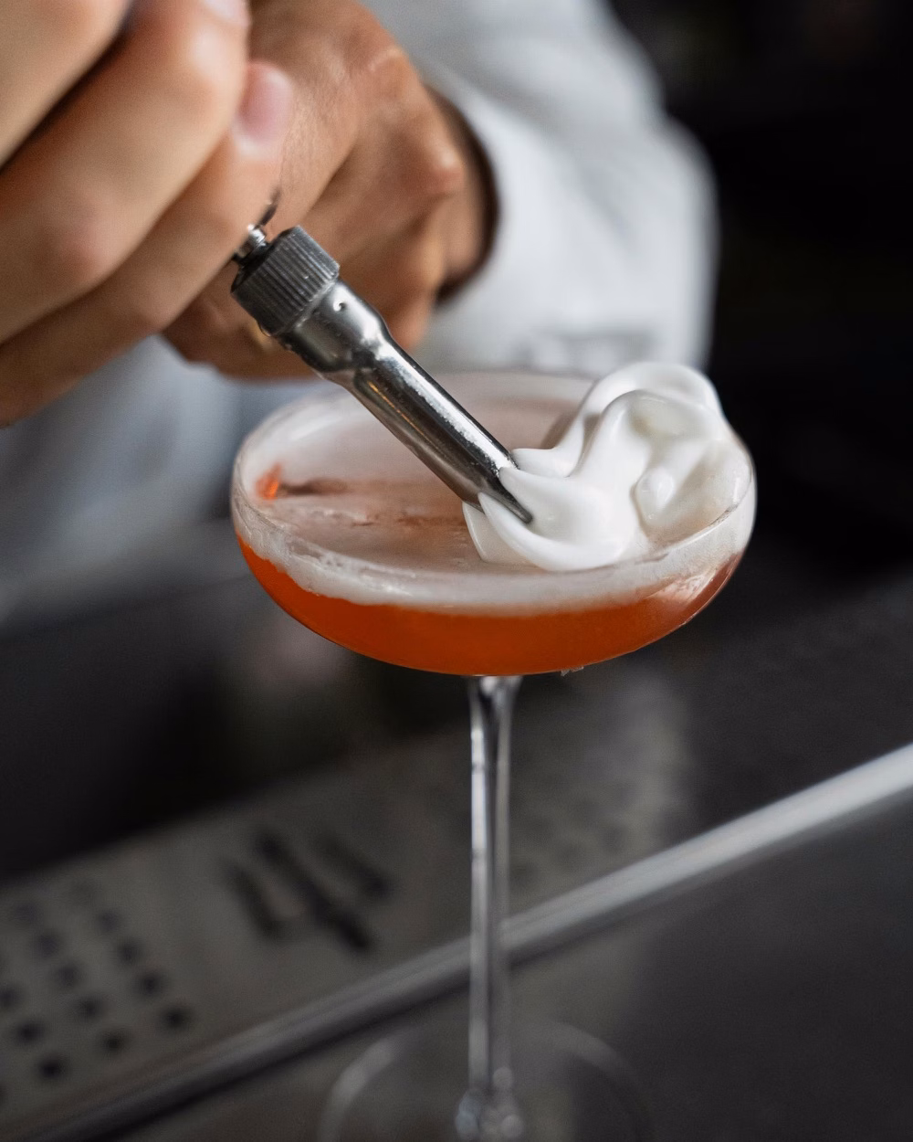 A bartender adds a white foam garnish to a pink cocktail in a stemmed glass