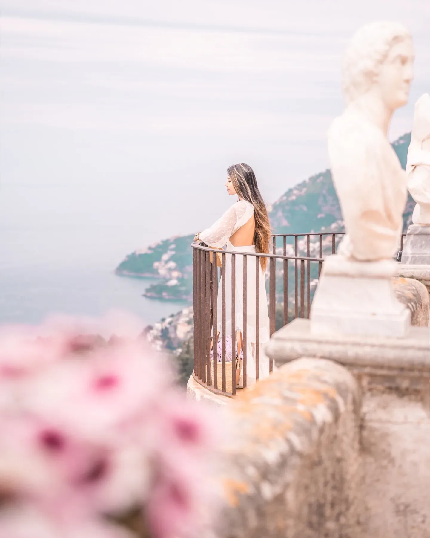 Woman in a flowing white dress gazes over a coastal cliff from a sunlit stone terrace with statues and soft pink flowers.