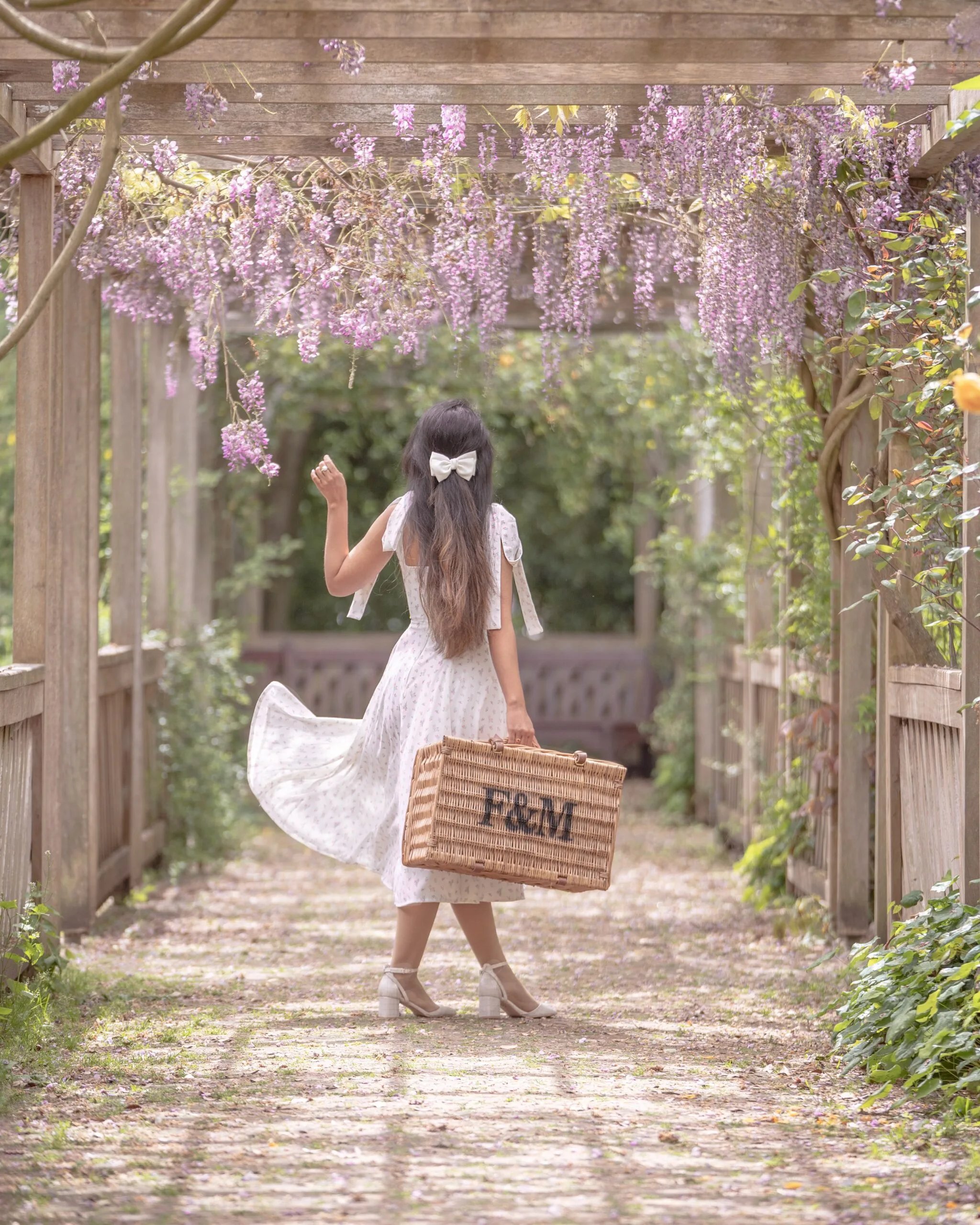 Woman in a white dress carrying a woven picnic basket beneath a pergola draped with hanging pink wisteria flowers.
