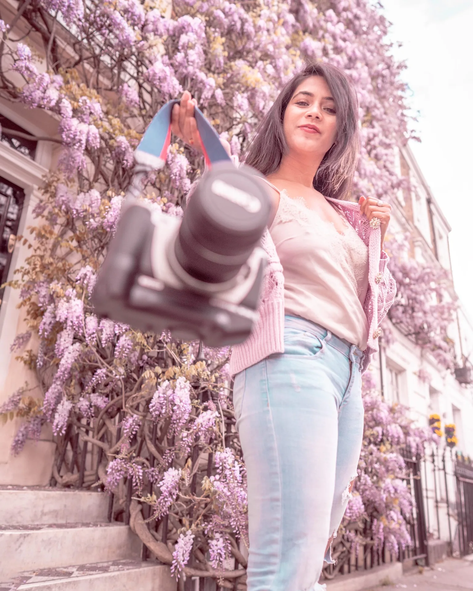Woman standing by a wisteria-covered townhouse offering a camera toward the viewer while smiling confidently.