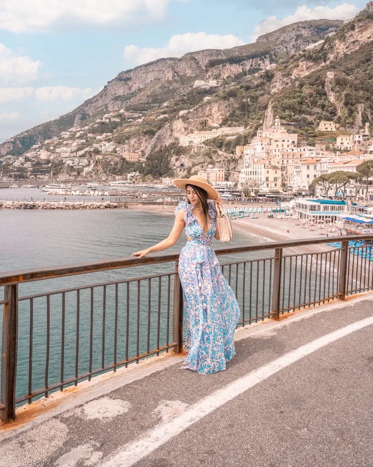 Woman in a floral maxi dress and straw hat leaning on a seaside promenade railing with Amalfi Coast cliffs and pastel buildings in the background.