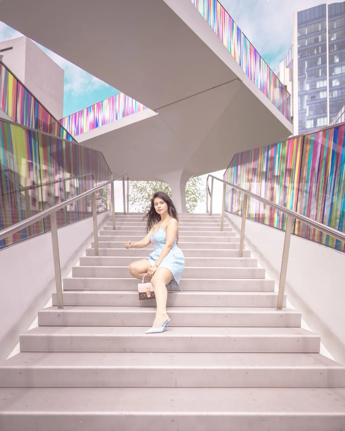 Young woman in a light blue dress sitting on wide concrete steps beneath colourful glass panels in a modern urban plaza