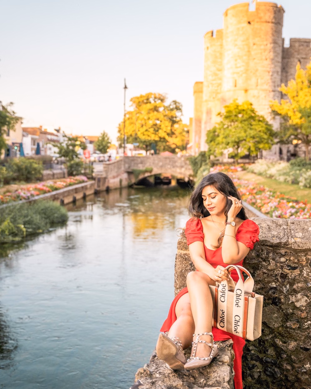 Woman in a red dress sits by a canal beside a historic stone tower and bridge at sunset