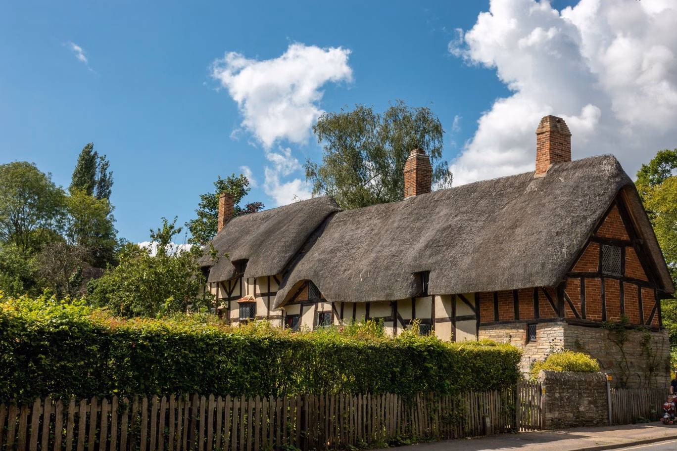 Thatched cottage with brick chimneys beside a hedge and wooden fence under a blue sky
