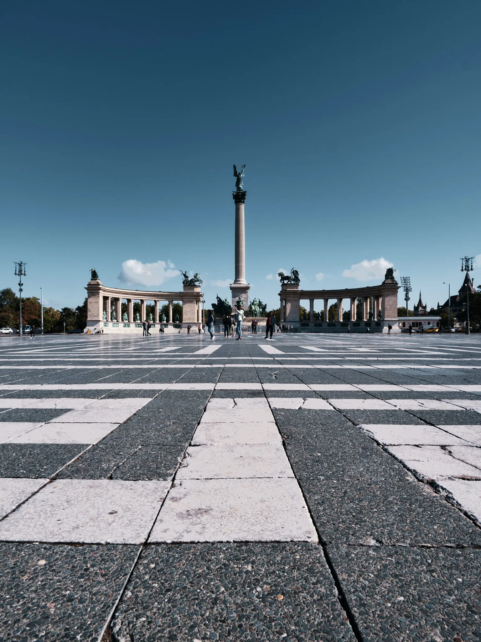 Wide low-angle view of Heroes' Square in Budapest with its central column, semicircular colonnades and checkered pavement under a clear sky.
