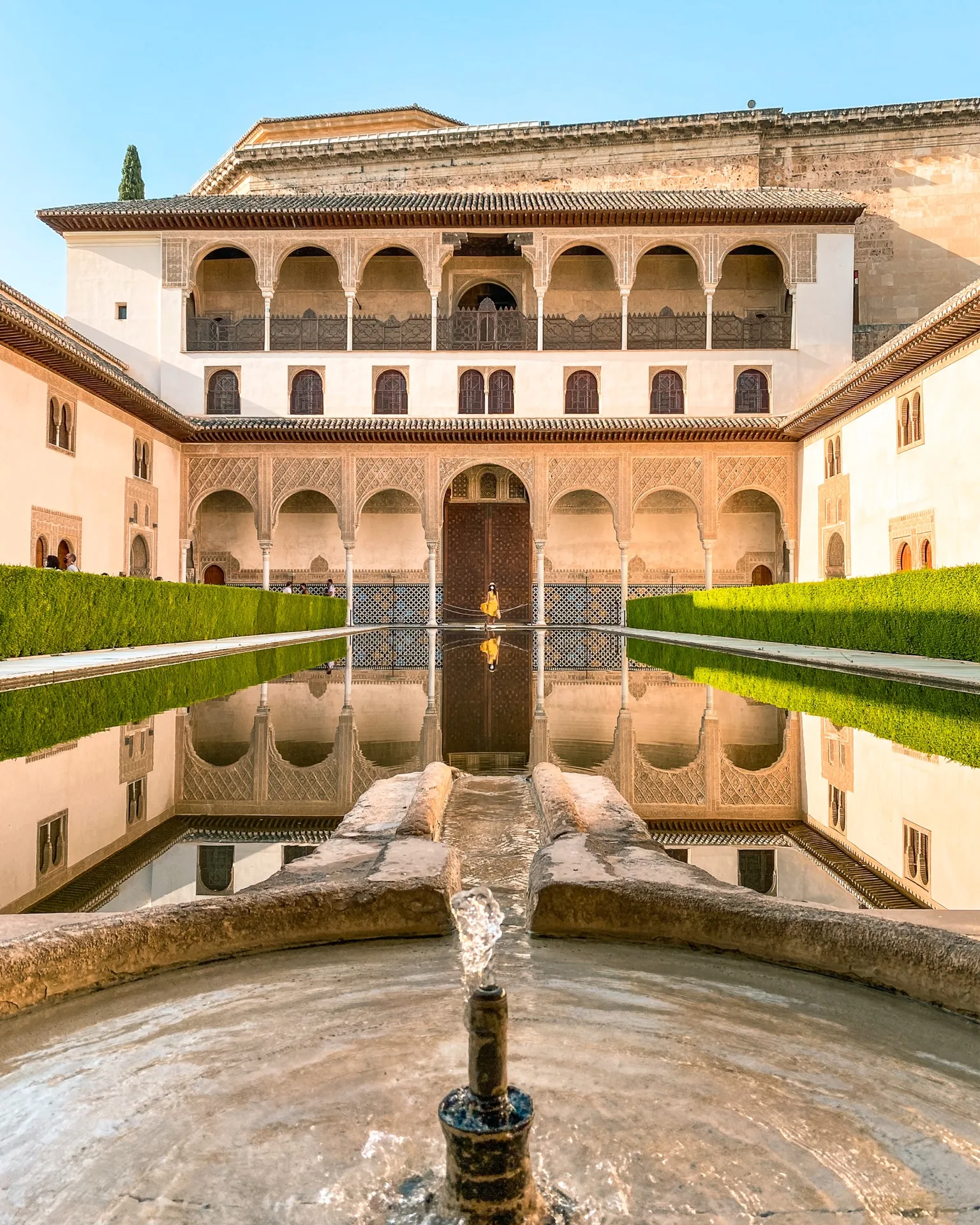 Ornate Moorish palace courtyard with central reflecting pool, symmetrical arches and a small fountain in the foreground.