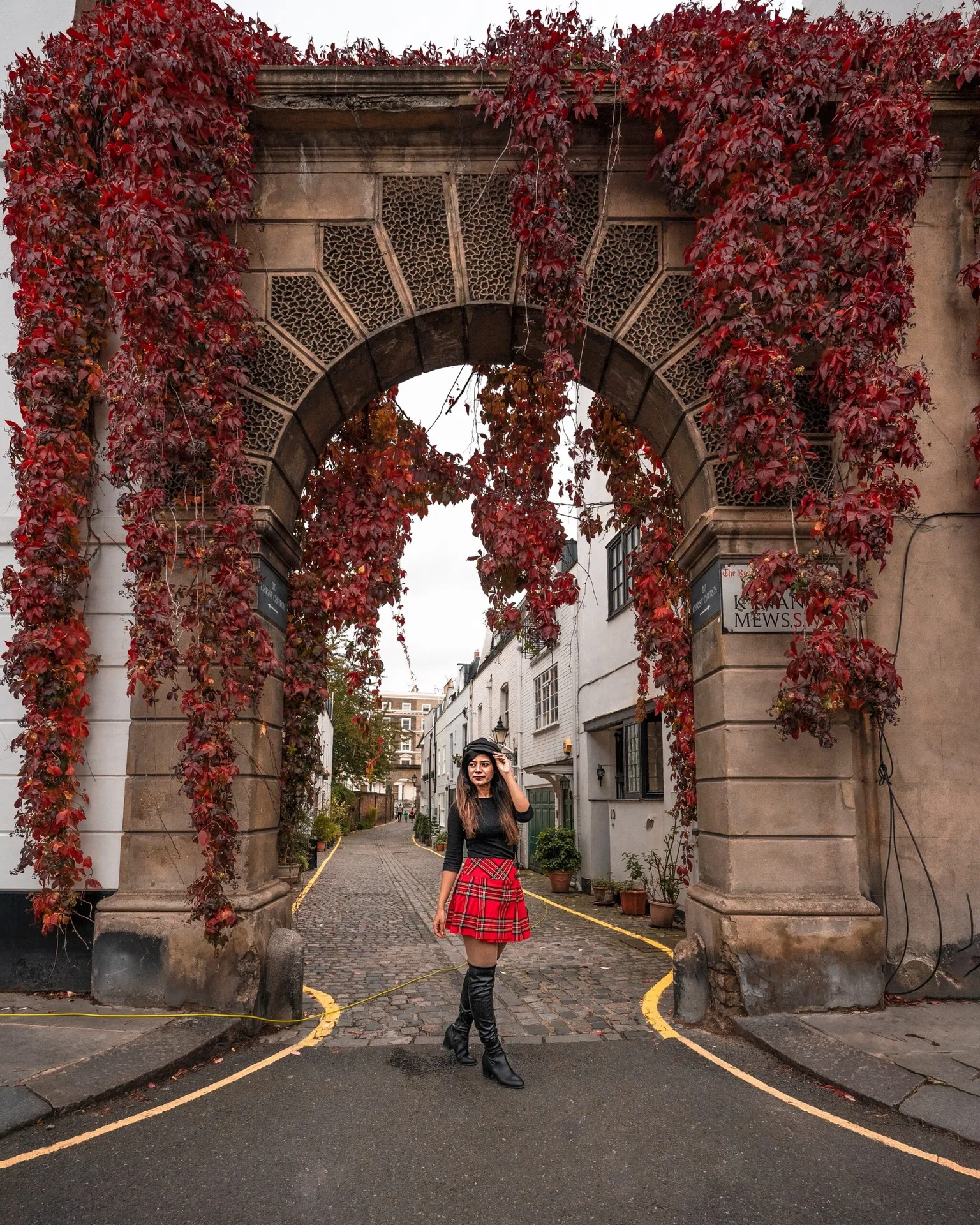 Woman in a red tartan skirt and black boots standing beneath a stone arch draped with red autumn ivy at the entrance to a cobbled mews.