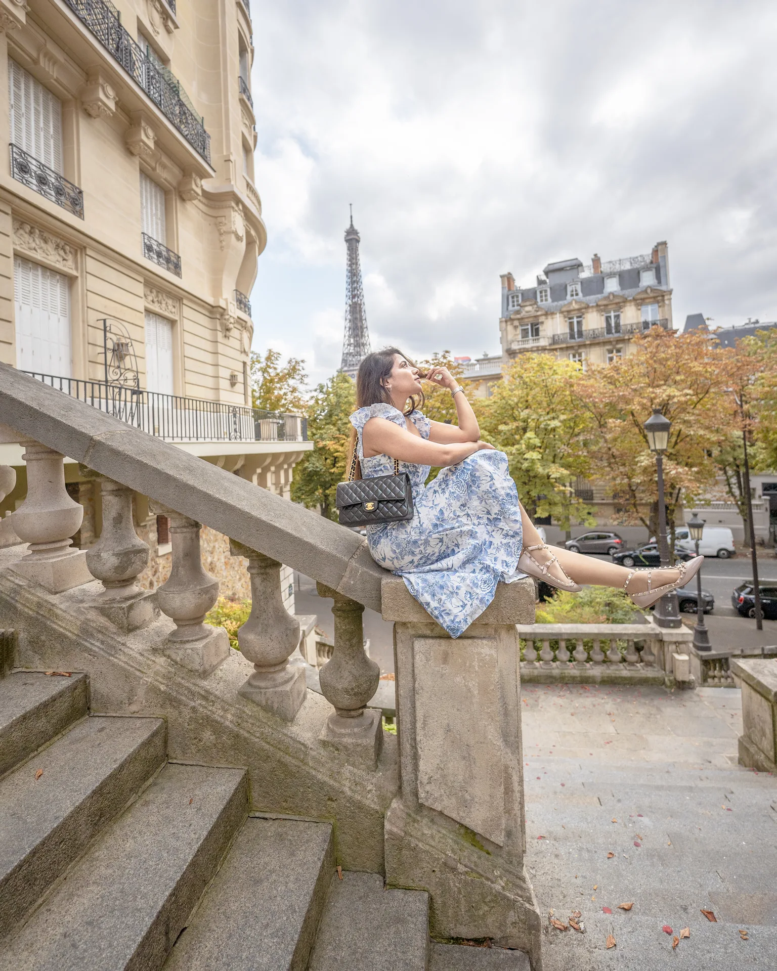 Woman in a blue floral dress sitting on stone balustrade with the Eiffel Tower and Parisian buildings in the background.