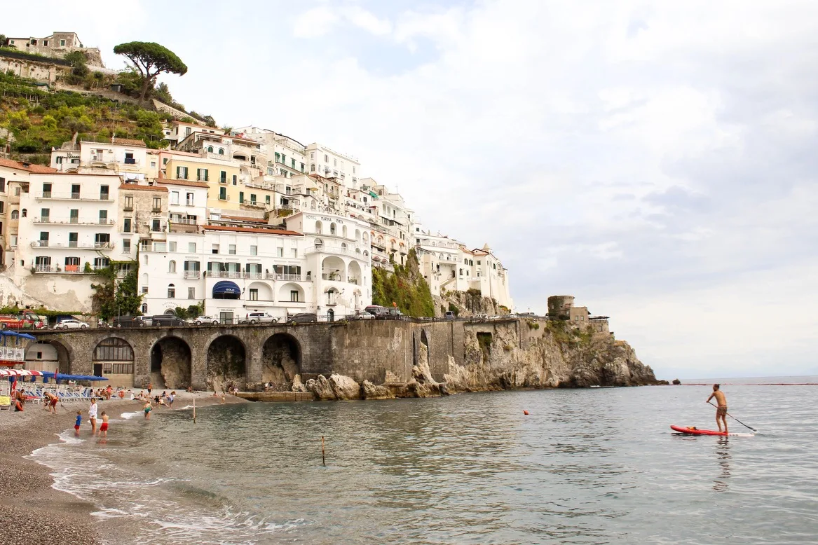 Sunlit cliffside town with white buildings cascading to a pebble beach where swimmers play and a paddleboarder glides on calm sea.