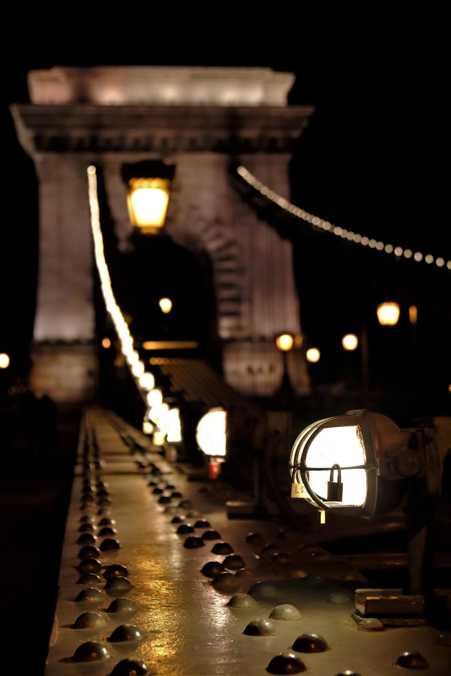 Night view along a riveted bridge railing lit by a row of warm lamps leading toward a softly illuminated stone arch.
