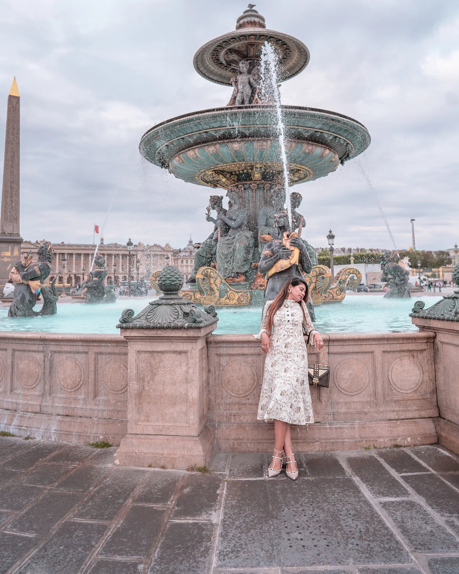 Woman in a floral dress leaning against the stone balustrade in front of an ornate fountain with sculptures and water jets at a grand city square