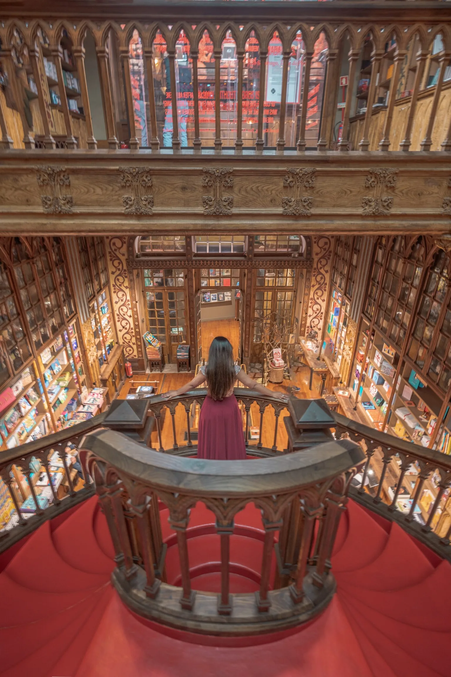 Woman in a red dress standing at the top of a curved wooden staircase overlooking an ornate, book-filled Victorian-style interior.