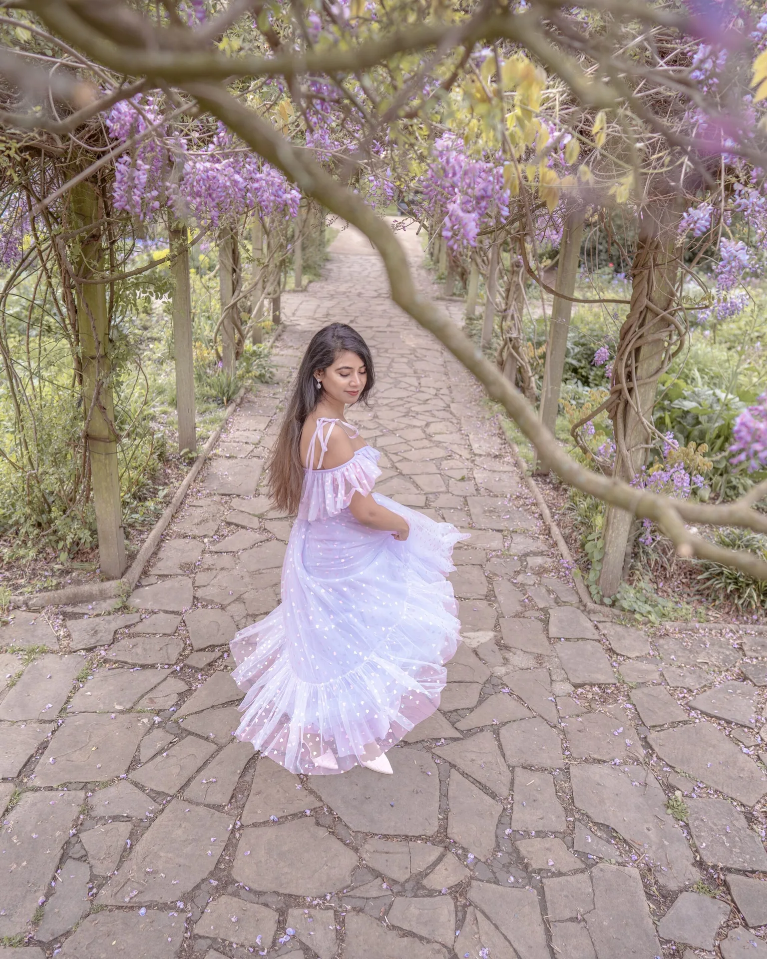 Woman in a flowing pastel dress twirling on a stone path beneath a pergola draped with blooming purple wisteria.