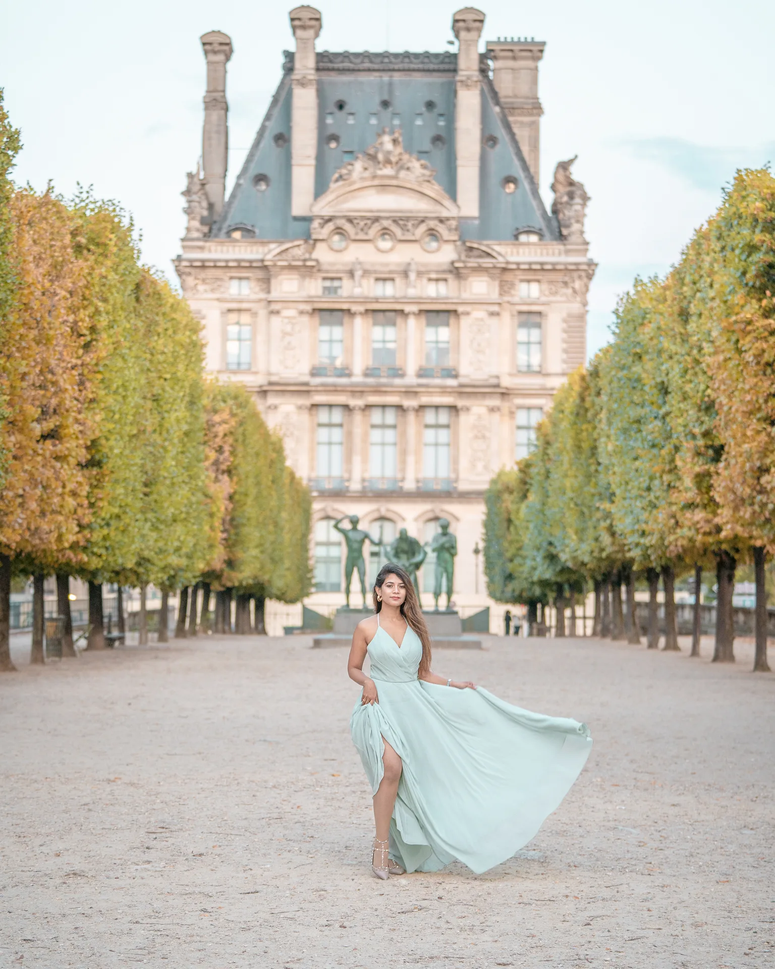 Woman in a flowing pale green dress posing on a tree‑lined path with a grand historic Louvre building in the background.