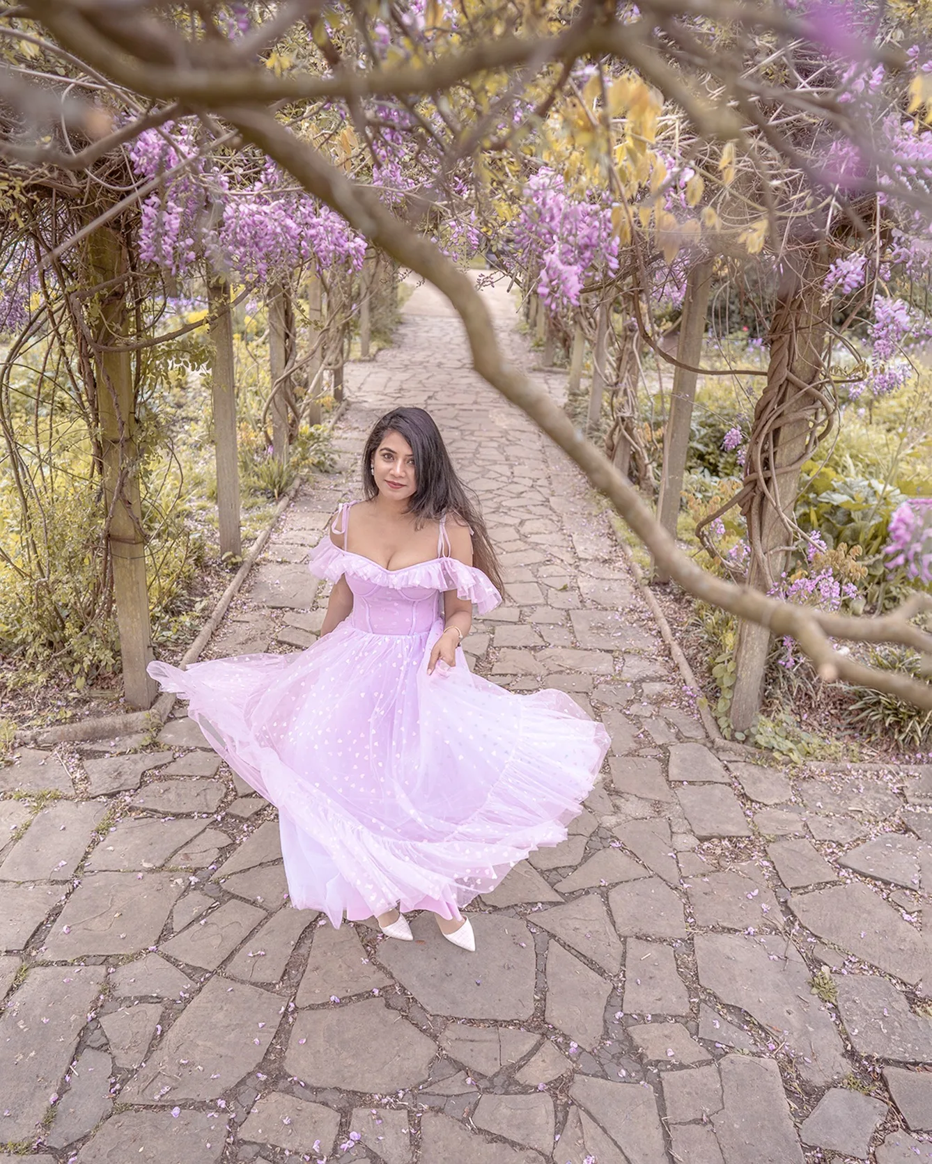 Woman in a flowing lilac dress twirling on a stone path beneath a wisteria-covered wooden pergola.