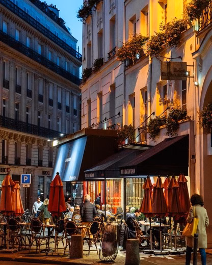 A lively evening café street with outdoor seating, red umbrellas, and warm lights between tall city buildings