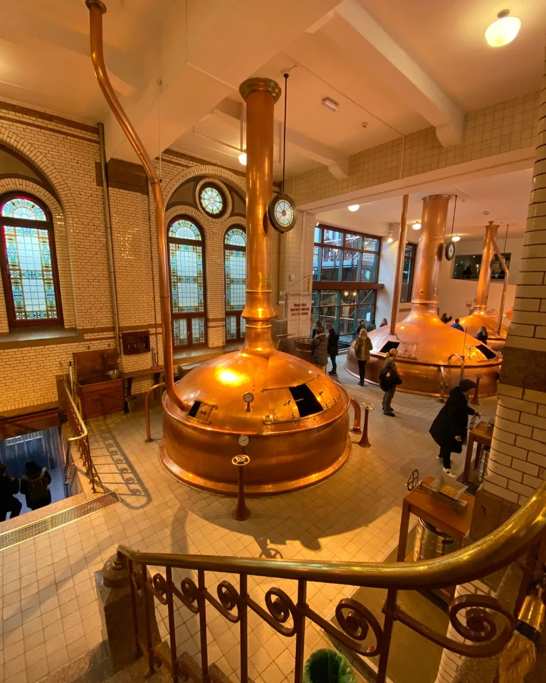 Visitors touring a historic brewery room with large polished copper brew kettles, tiled floors and stained-glass windows.