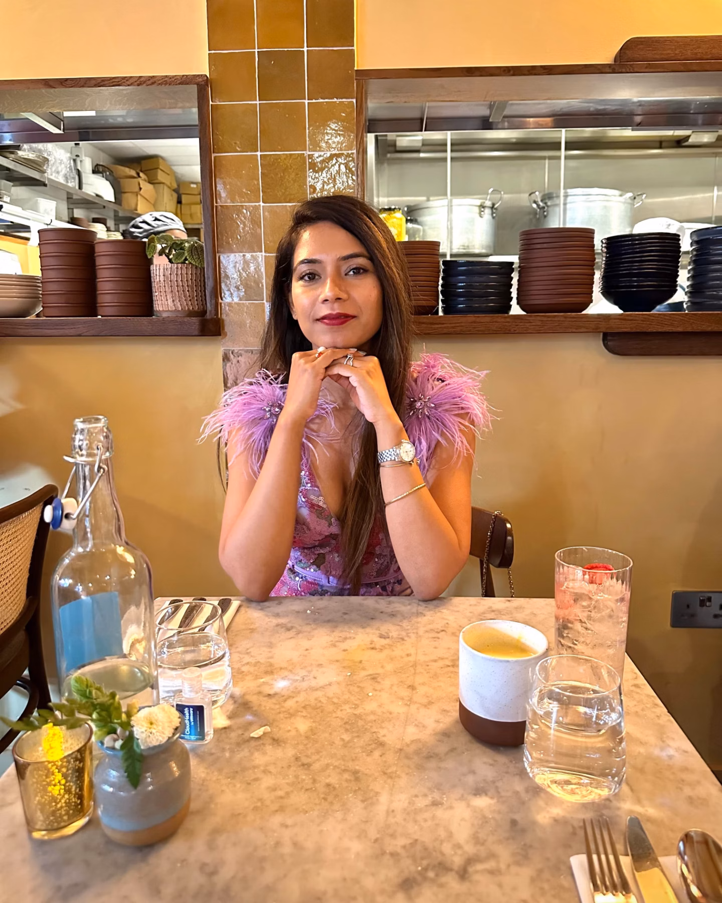 A woman in a lilac dress sits at a café table with drinks, smiling at the camera