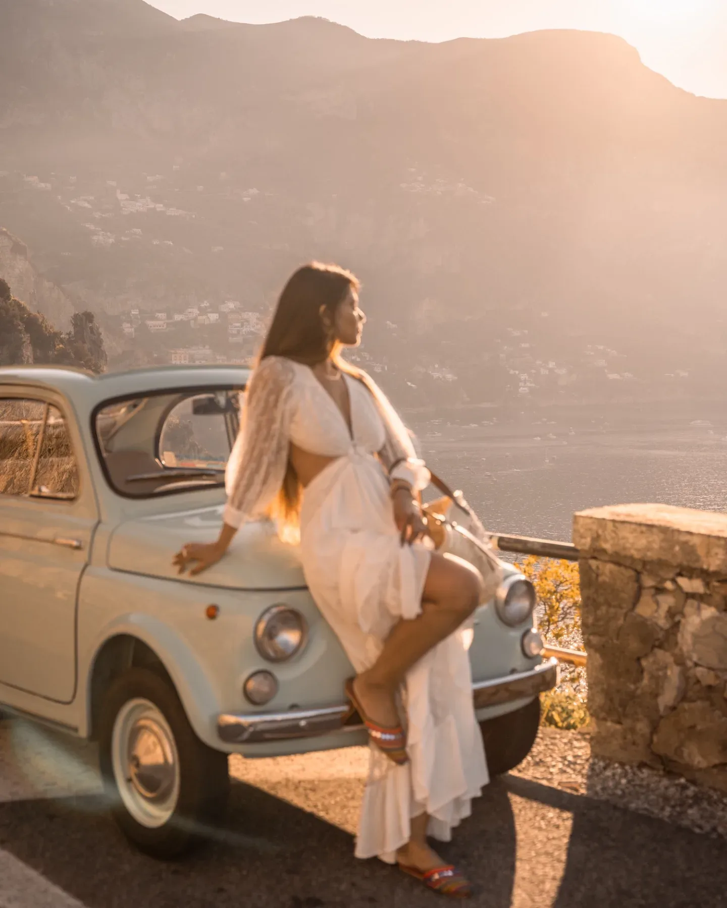 Best Instagram spots Amalfi Coast - Woman in a white dress leaning on a vintage pale blue car, gazing at a sunlit coastal cliffside at golden hour.