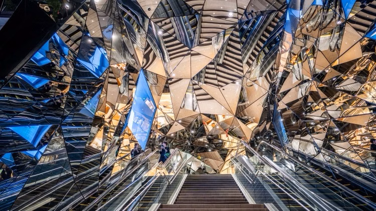 A mirrored, faceted interior with an escalator and stairs leading into a striking modern space