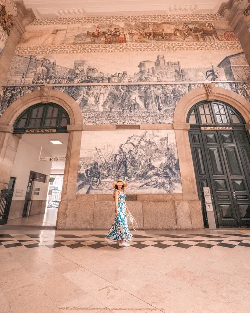Woman in a patterned dress and sunhat walking across a grand station hall beneath large blue-and-white tiled murals.