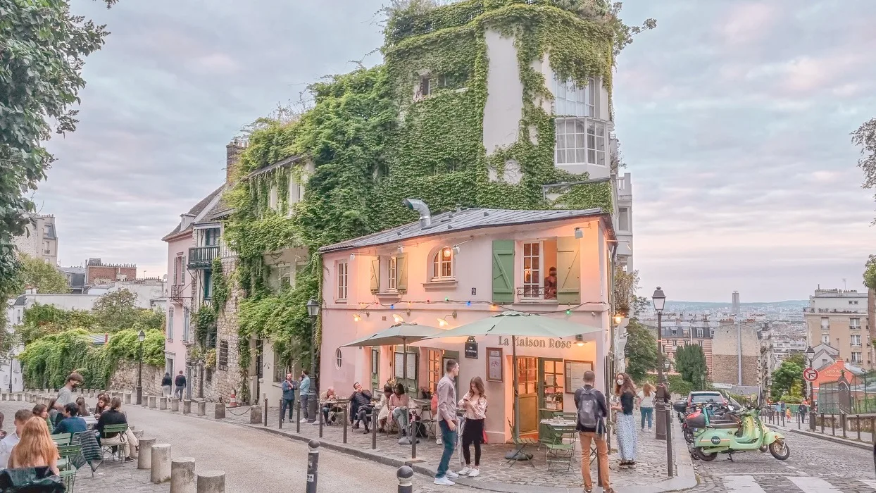 Quaint pink café covered in ivy with outdoor seating on a cobbled Montmartre street overlooking Paris at dusk.