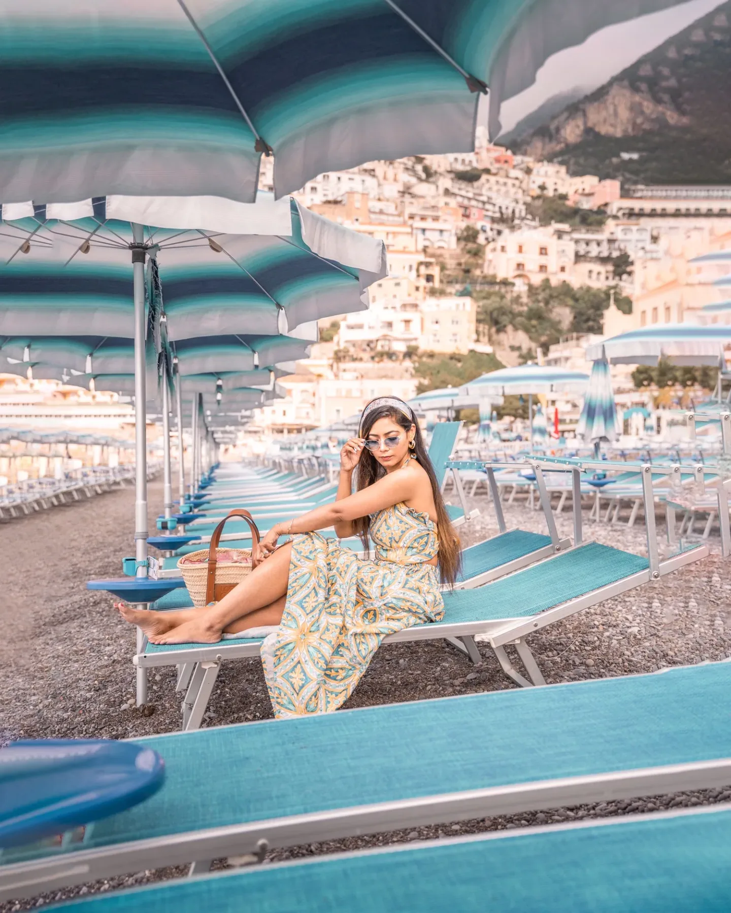 Woman in a patterned sundress and sunglasses lounging on a turquoise beach chair under striped umbrellas with a coastal town on the hillside behind her.