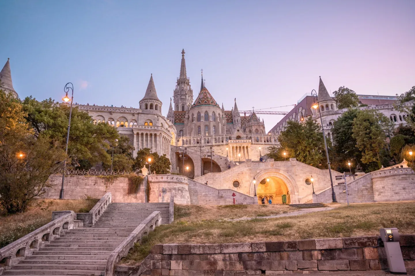 Ornate Fisherman's Bastion and Matthias Church atop a wide stone staircase, lit by warm evening lamps against a pastel sky.